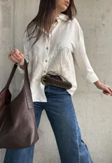 Camisa oversize blanca con botones frontales, cuello camisero y manga larga. Presenta grandes bordados en hilo beige con diseño de hojas o abanicos en la parte inferior frontal.
