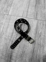 Cinto negro de cuero sintético con apliques metálicos plateados y hebilla metálica dorada.