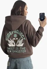 Hoodie de algodón de alto gramaje en color marrón, con capucha y bolsillo frontal. Presenta un diseño gráfico bordado en la espalda con dos cisnes y la frase 'Sweet is the New Gangster', con detalles en acabado aterciopelado.