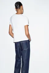 Camiseta de algodón color marrón oscuro, de corte regular, cuello redondo y manga corta. Presenta una estampación metalizada dorada en el parte frontal con texto "REYKJAVIK ART GALLERY" y "ABSTRACT MOVEMENTS Reykjavik, Iceland, 1975".