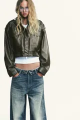 Cazadora corta de efecto piel color marrón oscuro, con cuello subido, manga larga con trabillas en los hombros, bolsillos con solapa en el delantero y cierre frontal con cremallera y botones a presión ocultos por solapa.