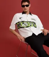 Camisa blanca oversized de tricolina con cuello negro contrastante, mangas cortas y estampado con la palabra "Hustle" en verde y negro.