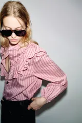 Camisa de manga larga con cuello subido, cierre frontal con botones y detalle de volantes en el pecho. Presenta un diseño a rayas verticales y puños elásticos.