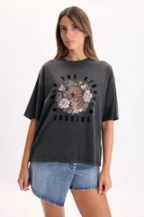 Camiseta boxy de manga corta color gris oscuro con efecto gastado, cuello redondo y estampa frontal que incluye un tigre rodeado de rosas y la frase "Only the strong survive".