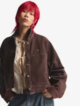 Chaqueta de lona de algodón color marrón con efecto lavado, corte holgado, cuello camisero y cierre frontal con botones metálicos. Presenta el logo de Adidas en dorado en el pecho y detalles de costuras en las mangas.