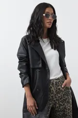 Trench coat de símil cuero color marrón oscuro, con solapas amplias, cierre frontal con botones, cinturón ajustable en la cintura y mangas con tiras ajustables. Presenta un largo por debajo de la rodilla y bolsillos laterales.