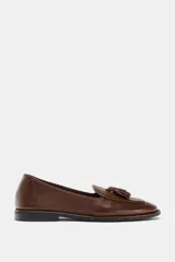Mocasín de cuero color marrón con detalle de borlas en el empeine y perforaciones decorativas tipo brogue en la puntera y el talón.