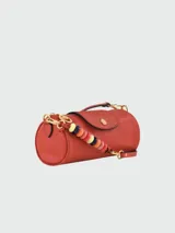 Cartera tipo crossbody de cuero color terracota, con forma cilíndrica, solapa frontal con cierre de botón a presión metálico y correa ajustable. Incluye un colgante decorativo de cuentas coloridas en uno de los laterales.