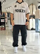 Camiseta deportiva blanca de manga corta con rayas verticales negras, cuello polo en V de color negro con ribete blanco, y puños negros. Cuenta con estampado frontal con la palabra "TOKIO" en grande, además de detalles gráficos y texto en el pecho.
