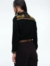 Camisa de color crudo con detalles de bordados en tonos mostaza y marrón en el cuello, hombros, bolsillos frontales y puños. Presenta cierre frontal con botones y dos bolsillos tipo cargo en el pecho.