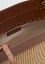Bolso shopper grande estilo capazo, confeccionado en tejido texturizado efecto rafia en tono beige, combinado con detalles de cuero sintético marrón en los bordes y las asas dobles de hombro. Cuenta con cierre de cremallera superior y un charm colgante decorativo.