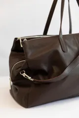 Bolso de viaje tipo weekender de cuero vacuno color marrón chocolate, con doble asa de mano y correa bandolera ajustable y extraíble. Tiene cierre superior con cremallera y múltiples bolsillos internos.