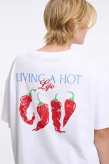 Remera de corte oversized en color blanco, con cuello redondo y mangas cortas. Presenta una estampa central con la frase 'Living a hot life' y un diseño de ajíes rojos.