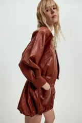 Chaqueta corta estilo bomber de Zara, confeccionada en material efecto piel color marrón rojizo. Presenta cuello redondo, hombreras, mangas abullonadas con puños elásticos y cierre frontal con cremallera oculta y botones a presión. Incluye bolsillos delanteros de vivo.