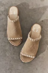 Sandalias tipo mule de gamuza color tostado, con detalle de costura artesanal en el borde superior y en la puntera abierta. Poseen suela plana con textura y plantilla con logo grabado.