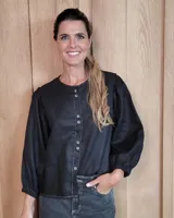 Camisa negra de corte holgado con cuello redondo, cierre frontal con botones y mangas largas abullonadas con puños elásticos. Presenta costuras visibles en contraste.