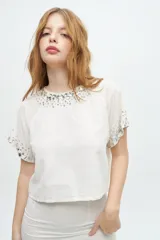 Blusa de manga corta color crudo, con escote a la base decorado con pedrería y cristales bordados. Presenta un diseño de corte recto y holgado con detalles de pedrería también en los bordes de las mangas.