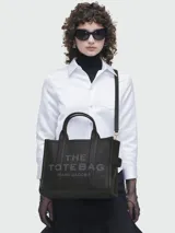 Cartera tote de cuero granulado negro con diseño estructurado. Presenta el logo 'THE TOTE BAG' grabado en el frente, doble asa superior y correa de hombro ajustable y extraíble.