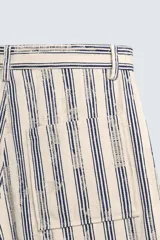 Bermuda de hombre color beige con estampado de rayas verticales azules con efecto desgastado. Corte relaxed fit, con cierre frontal de cremallera y botón, bolsillos delanteros y bolsillos traseros de plastrón.