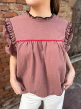 Blusa color chocolate con mangas cortas abullonadas, cuello redondo con volados y detalles de costura en zigzag de color rojo en el pecho y volados oscuros en los hombros.