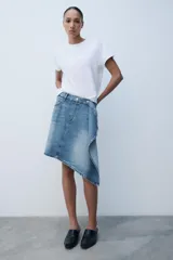 Falda de tiro medio confeccionada en denim, con trabillas, cinco bolsillos, cierre cruzado con botones y bajo acabado asimétrico.