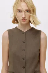 Chaleco largo de vestir color marrón oscuro, con cuello en V, cierre frontal de botones y bolsillos ribeteados. Presenta un corte entallado y abertura en la parte trasera.