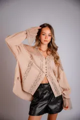Camisa holgada color beige, con mangas largas acampanadas y detalles de encaje en el canesú, hombros y puños. Presenta un escote en V con botones frontales.