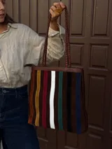 Cartera tipo tote de cuero con diseño de franjas verticales multicolores en tonos tierra, rojo y azul. Cuenta con correas trenzadas de cuero y cierre superior.