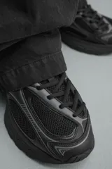 Zapatilla deportiva tipo running de color negro, confeccionada en una combinación de piezas de malla y material sintético. Presenta un diseño con cordonera curvada y una suela de volumen con diseño irregular.