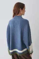 Buzo tejido de silueta amplia con cuello alto, mangas largas y puños en contraste. Presenta un diseño con franjas horizontales en el ruedo y detalles de tejido trenzado en las mangas.