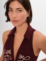 Chaleco de pana color bordó, ajustado al cuerpo, con pinzas y elásticos en la espalda baja. Presenta un largo irregular, cuello clásico camisero y cierre frontal con ganchos peleteros. Tiene bordados decorativos en color crema en el frente.