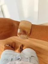 Brazalete tipo cuff de metal dorado con diseño de relieve ondulado y acabado brillante. Es de diseño abierto y ajustable.
