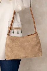 Cartera de hombro confeccionada en gamuza color beige, con detalles en cuero sintético marrón en las manijas y bordes. Presenta un diseño minimalista y funcional con cierre superior.