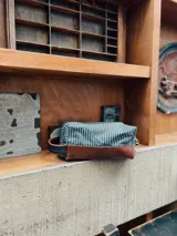 Neceser de diseño rectangular con cuerpo de tela a rayas verticales blancas y negras, y base reforzada de cuero marrón. Cuenta con una manija lateral de cuero negro para facilitar su transporte.