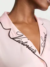 Pijama corto de satén color rosado con detalles de ribetes negros, compuesto por una camisa de manga corta abotonada con bolsillo en el pecho y el logotipo bordado en el cuello, y un short con cintura elástica.