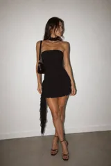 Vestido corto negro de estilo strapless con diseño ajustado al cuerpo y falda con volados en capas. Incluye un detalle de cinta tipo choker en el cuello y una caída lateral de tela transparente.