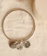 Pulsera rígida plateada tipo esclava, elaborada en alpaca, con tres dijes redondos colgantes. Uno de los dijes tiene grabada la letra 'M'.