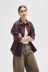 Sobrecamisa de cuero sintético en tono bordó oscuro, con cuello clásico, cierre frontal oculto y un bolsillo aplicado en el pecho. Presenta un corte holgado con mangas largas y puños rectos.