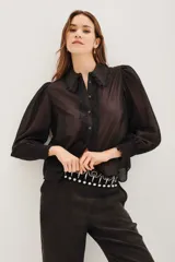 Camisa negra de manga larga con diseño de hombros abullonados y puños elásticos. Presenta un cuello clásico y una tapeta frontal con botones, decorada con detalles de encaje floral a lo largo de la abertura.
