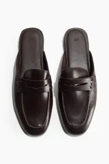Mules estilo mocasín color marrón oscuro, con diseño destalonado, punta redondeada y tira frontal característica del modelo penny loafer.