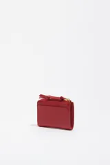 Monedero o tarjetero pequeño de cuero rojo, con cierre de cremallera superior y un tirador de tela a juego. Presenta un bolsillo exterior frontal y el logo de la marca grabado en dorado.