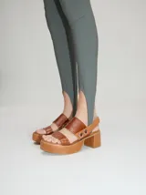 Sandalias de plataforma con taco cuadrado, confeccionadas en cuero marrón con tiras de charol a juego. Presentan una correa trasera con elástico y detalle de doble argolla metálica en los laterales.