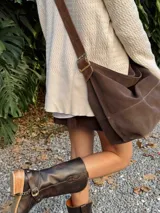Bolso tipo bandolera de gran tamaño, confeccionado en gamuza color marrón. Presenta una correa ancha ajustable con hebilla metálica y costuras visibles en el cuerpo del bolso.