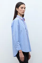 Camisa de corte clásico con estampado de rayas verticales en tonos celestes y blancos. Presenta un detalle de bordado decorativo en color rojo a lo largo de la botonadura frontal y el cuello, mangas largas con puños abotonados y un bolsillo frontal en el pecho.
