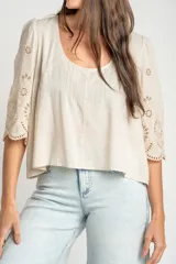 Blusa color arena de algodón peinado, con escote redondo, mangas cortas abullonadas con bordados suizos y detalles de alforcas en el frente.