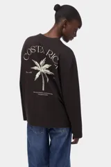 Remera de manga larga oversize confeccionada en punto de algodón suave, color marrón oscuro, con cuello redondo ribeteado, hombros caídos y pequeño estampado tipográfico en el pecho.