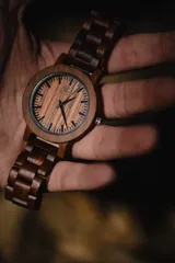 Reloj de pulsera con caja, esfera y malla de madera de sándalo rojo, con marcadores y manecillas negras. Presenta un diseño circular con corona lateral.