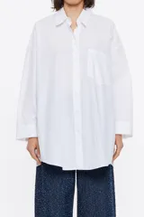 Camisa larga blanca de algodón, de corte recto, con cuello camisero, manga larga, puños con botones, cierre frontal de botonadura y bolsillo de plastrón en el pecho con logo en relieve.