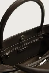 Bolso de mano estructurado en color marrón, confeccionado en una combinación de materiales que incluye gamuza en el cuerpo principal y cuero en la parte superior y asas. Presenta un detalle de cinturón decorativo con hebilla metálica plateada en la parte frontal, doble asa de mano y cierre principal con imán.