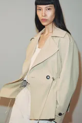 Trench corta de color beige con cuello de solapa y manga larga. Cuenta con bolsillos delanteros, cintura ajustable con cordones, bajo abullonado y cierre frontal cruzado con un botón visible. El interior está forrado con tela a cuadros.